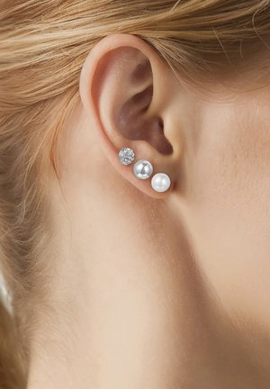 Close-up af en persons øre med tre ørestikker: en med klare krystaller, en sølvmetallic og en hvid perle.