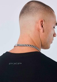 Hombre con corte de cabello corto al rape que lleva una camiseta negra con el texto "PXP" en la parte de atrás y un collar grueso de plata.