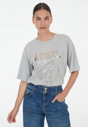 Frau mit braunem, zurückgebundenem Haar, die ein hellgraues AUREL-Grafik-T-Shirt und blaue, hoch taillierte Jeans trägt, mit einer Hand in der Tasche.