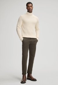 Maglione a coste color crema con collo alto abbinato a pantaloni chinos verde oliva. Il modello indossa stivaletti in pelle marrone scuro. Sfondo semplice, colori neutri.