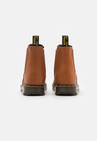 Brązowe skórzane botki chelsea o matowym wykończeniu, czarne gumowe podeszwy, żółte przeszycia oraz pętle z żółtym tekstem na czarnym tle.