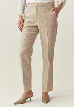 Žena v béžových kalhotách na míru s předními záhyby, bílé blůze a taupe nazouvacích botách na nízkém podpatku s špičatou špičkou, stojící s jednou rukou v kapse.