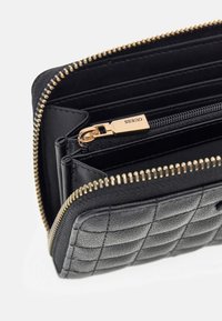 Cartera acolchada negra con cremallera dorada, múltiples ranuras para tarjetas y un compartimento central con cremallera etiquetado "GUESS."