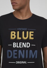 Melna kokvilnas T-kreklu ar vintage stila, daudzkrāsainu teksta dizainu: "BLUE," "BLEND," "DENIM," dzeltenā, baltā un zilā krāsā.