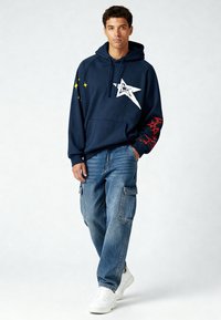 Sweat à capuche bleu marine avec graphique d'étoile blanche et accents rouges sur les manches, associé à un jean cargo délavé clair et des baskets blanches.