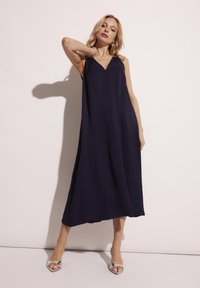 Granatowa sukienka bez rękawów z dekoltem w serek, wykonana z lekkiego materiału, z luźnym krojem. Modelka nosi srebrne sandały na obcasie.