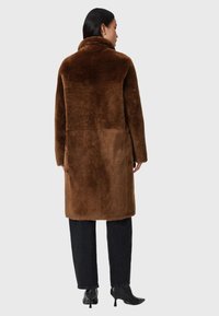 Cappotto in pelliccia artificiale marrone con collo alto, maniche lunghe e vestibilità rilassata, abbinato a jeans neri e stivali con tacco. Texture morbida, cuciture minime.