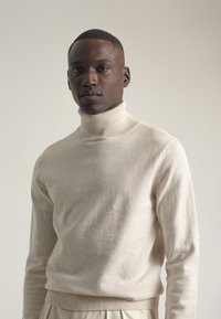 Hombre negro con cabello corto que lleva un suéter de cuello alto beige y pantalones color crema, contra un fondo claro y sencillo.