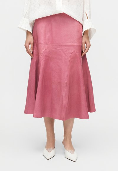 Ροζ δερμάτινη φούστα με φαρδιά γραμμή, με καμπύλες ραφές και διακριτή υφή, συνδυασμένη με λευκά παπούτσια μυτερά στη μύτη.