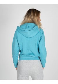 Felpa turchese con cappuccio, dal taglio rilassato e con polsini a coste. Tessuto in cotone morbido, vista dal retro, abbinata a pantaloni della tuta grigi chiari.