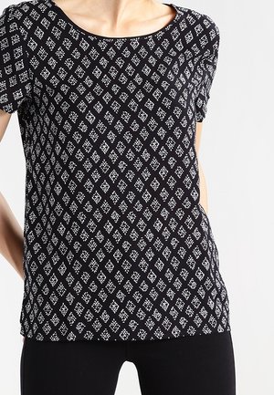 Kobieta ubrana w czarny top z krótkim rękawem z białym wzorem diamentów i kropek, połączony z czarnymi spodniami, na jasnym tle.