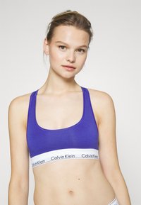 Fialový športový podprsenkový top z bavlneného zmesového materiálu s širokým bielym pásom s čiernym logom "Calvin Klein", zaobleným výstrihom a dizajnom s prekríženými ramienkami.