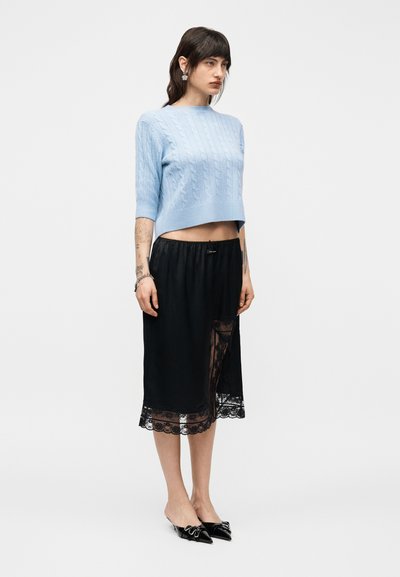 Femme debout portant un pull court en maille torsadée bleu clair, une jupe midi noire avec bordure en dentelle, et des ballerines noires pointues avec un détail nœud.