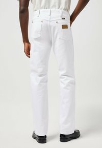 Jean en denim blanc avec une coupe droite, poche arrière en patch et accents en rivets cuivrés. Comprend une étiquette de marque en cuir et une texture subtile.