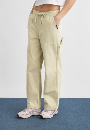 Osoba oblečená v béžových nohaviciach so šnúrkou v páse a bočnými vreckami, obutá v svetloružových teniskách, stojaca na modrom koberci pred jednoduchým bielym pozadím.