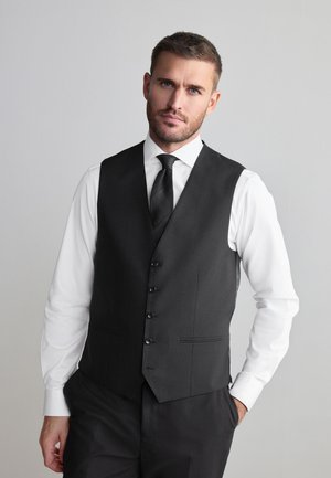 Uomo con capelli corti e barba, indossa camicia bianca, cravatta nera e gilet nero, in piedi con una mano in tasca davanti a uno sfondo grigio.