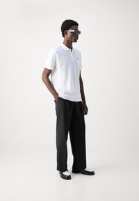 Hvit polo skjorte med korte ermer og krage, kombinert med løse svarte bukser og svarte sko med hvite detaljer. Modell som bruker solbriller.