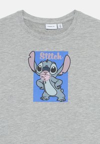 Szürke póló, amelyen egy rajzfilmfigura, Stitch áll a kezeire támaszkodva, rózsaszín rágógumibuborékot fújva, kék háttér előtt.