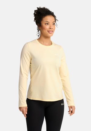 Donna con capelli ricci che indossa una maglia a maniche lunghe giallo chiaro e leggings neri in piedi davanti a uno sfondo semplice.