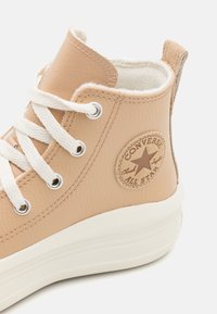 Zapatilla alta de cuero beige con cordones blancos, un interior suave y una suela de goma marrón; presenta un parche circular de la marca en el costado.