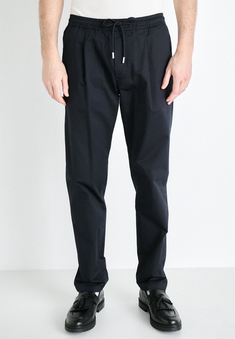 Homme portant un pantalon à cordon noir et des mocassins à pampilles noirs, debout sur un fond blanc.