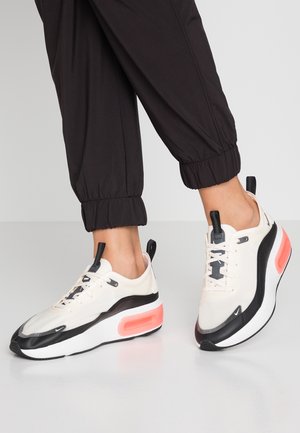 Pieds portant des baskets compensées blanches, noires et roses avec des languettes noires, associées à un pantalon de jogging noir retroussé, sur un sol gris clair.