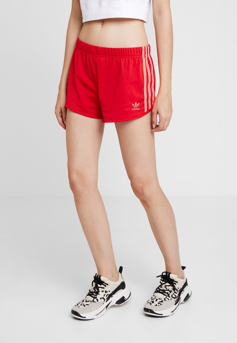 Žena v červených sportovních šortkách Adidas s růžovými pruhy a bílých teniskách s černými tkaničkami, stojící před jednoduchým bílým pozadím.