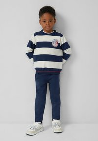 Jongen staat met zijn handen achter zijn rug, draagt een marineblauwe en witte gestreepte trui, marineblauwe broek en witte sneakers tegen een effen achtergrond.