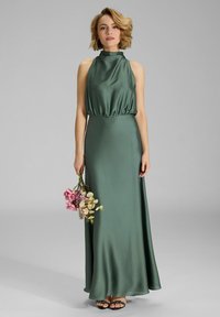 Dunkelgrünes Satinabendkleid mit Halterneck, elastischem Taillenbund und fließendem Rock. Das Model hält einen Blumenstrauß. Sichtbare schwarze Riemchensandaletten.