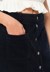 Marineblå kordfløjlskørt med frontlukning med knapper, sidelommer og lodret ribbet tekstur. Stylet med en hvid flæsebluse.