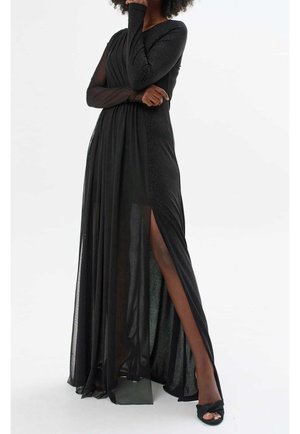 Kvinde i lang sort kjole med gennemskinneligt stof, høj slids på venstre ben og lange ærmer, poser med hånden nær ansigtet og armene krydsede.