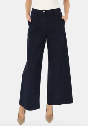 Kobieta w granatowych szerokich spodniach i beżowych szpilkach z ostrym noskiem, z rękami w kieszeniach, stoi na jednolitym tle.