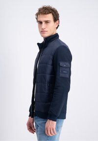Man wearing a navy quilted jacket with zippered pockets and a flap pocket on the sleeve, paired with light blue jeans, standing sideways.