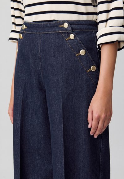 Pantalones de mezclilla oscura con un diseño de pierna ancha, que presentan una cintura alta, botones dorados a lo largo del lado izquierdo y detalles de costura visibles.