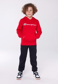 Red hoodie with white "Champion" logo, front pocket, paired with black sweatpants and black sneakers with white accents.