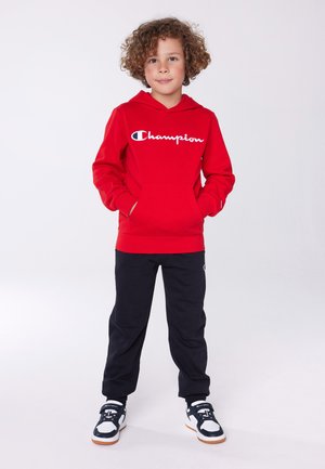 Sudadera roja con el logo blanco de "Champion", bolsillo frontal, combinada con pantalones de chándal negros y zapatillas negras con acentos blancos.