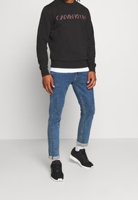 Felpa nera in cotone con logo "CALVIN KLEIN", abbinata a jeans in denim blu e scarpe da ginnastica nere, con le maniche arrotolate.