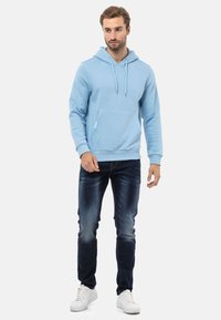 Helles blaues Hoodie aus Baumwolle, ausgestattet mit einer Fronttasche, Kapuze mit Kordelzug und gerippten Bündchen, kombiniert mit dunkelblauen Jeans und weißen Sneakers.