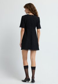 Musta tweed-mekko lyhyillä hihoilla, helma frillayksityiskohdilla ja takana vetoketju. Yhdistetty mustiin polviin ulottuviin sukkahousuihin ja teräväkärkisiin korkokenkiin.