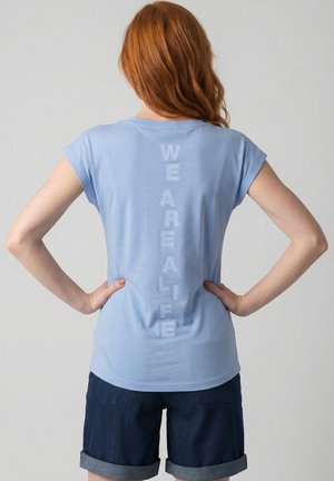 Frau mit roten Haaren steht mit Händen in der Hüfte, trägt ein hellblaues T-Shirt mit vertikalem Schriftzug "WE ARE ALIFE" und dunkel umgekrempelte Shorts.
