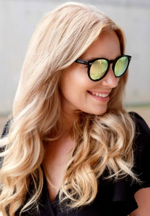 Černé sluneční brýle s kulatými obrubami a zelenými skly nosí osoba s dlouhými, vlnitými blond vlasy, které jsou částečně zakryty brýlemi.