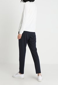 Persona con un top bianco a maniche lunghe, pantaloni neri e sneaker bianchi, in piedi con le spalle rivolte verso chi guarda, su uno sfondo neutro.