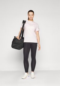Lys rosa Champion t-skjorte med logo, svarte leggings og hvite joggesko. Svart skulderveske med teksturert detalj. Nøytral bakgrunn.