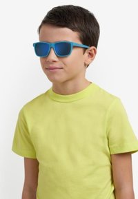Occhiali da sole blu con lenti scure e finitura liscia, indossati da un bambino in una maglietta gialla brillante, in piedi su uno sfondo chiaro.