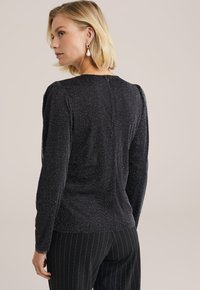 Top nero a maniche lunghe con tessuto texturizzato e luccicante, caratterizzato da spalle a sbuffo e chiusura posteriore. Indossato con pantaloni a righe.