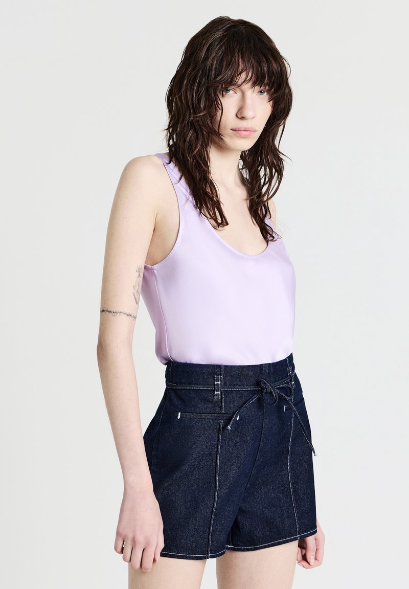 Giovane con capelli scuri all'altezza delle spalle, indossa una maglietta senza maniche color lavanda e pantaloni shorts in denim scuro a vita alta con cintura annodata.