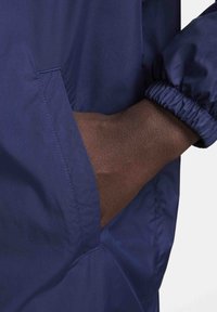 Marinblå vindjacka med elastiska ärmslut, med en sidficka och slät yta. Synlig sömnad längs sömmarna ger detalj.