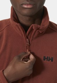 Polarowa kurtka w kolorze wypalanej pomarańczy z wysokim kołnierzem, pół-zamkiem z przodu oraz czarnym logo na klatce piersiowej. Ręka trzymająca suwak.