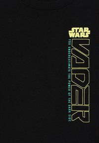 Svart bomull t-shirt med gul och teal text, inklusive "STAR WARS" och "VADER" med ett citat om den mörka sidan, vertikalt justerad.