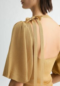 Maglia dorata a maglia con maniche a sbuffo, schiena aperta e dettaglio con fiocco. La texture liscia mette in risalto il design aderente.